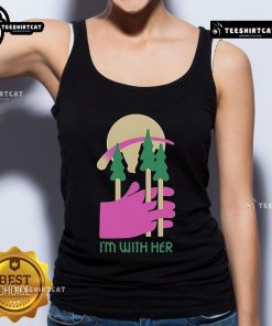Alt Text: Woman wearing a 'Nice, I'm With Her' tank top surrounded by three trees in a sunny outdoor setting.