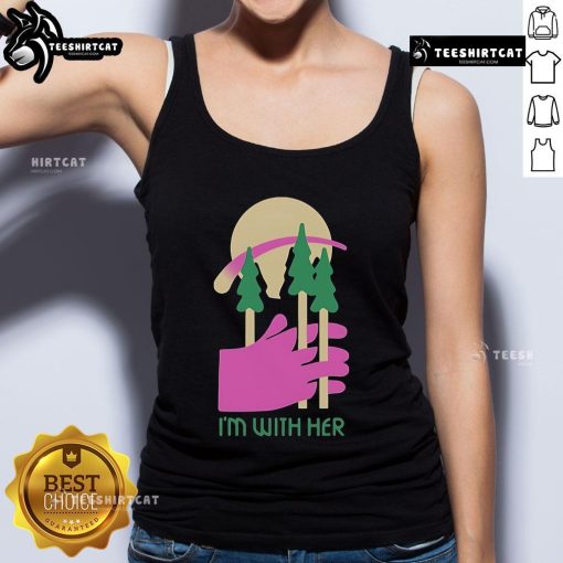 Alt Text: Woman wearing a 'Nice, I'm With Her' tank top surrounded by three trees in a sunny outdoor setting.