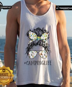 Mom in a messy bun wearing a tank top enjoying camping life surrounded by nature and outdoor gear.