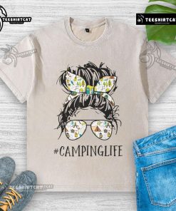 Mom enjoying a pretty camping life in a messy bun, wearing a washed T-shirt, surrounded by nature and camping gear.