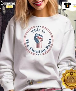 Charlie Kirk fist raising in front of a US flag, wearing a 'Top-This Is The Turning Point' sweatshirt, showcasing patriotism.
