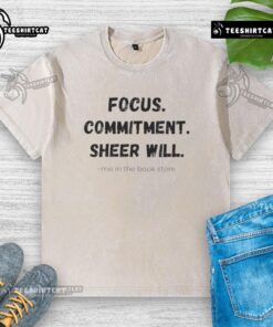 Official Focus Commitment Sheer Will Me in the Book Store washed t-shirt displayed on a wooden shelf with books.