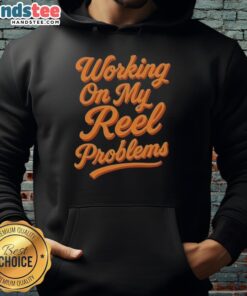 Person wearing a 'Happy Working On My Reel Problems' fishing hoodie, smiling while holding fishing gear outdoors.