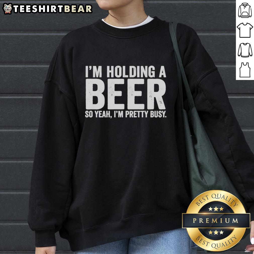 awesome-im-holding-a-beer-so-year-im-pretty-busy-sweatshirt Person wearing an 'Awesome I'm Holding A Beer' sweatshirt, smiling and enjoying a casual moment with a drink.