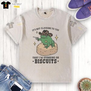 Alt Text: Cute frog cowboy holding a pistol in a washed t-shirt, standing on biscuits in a whimsical outdoor setting.